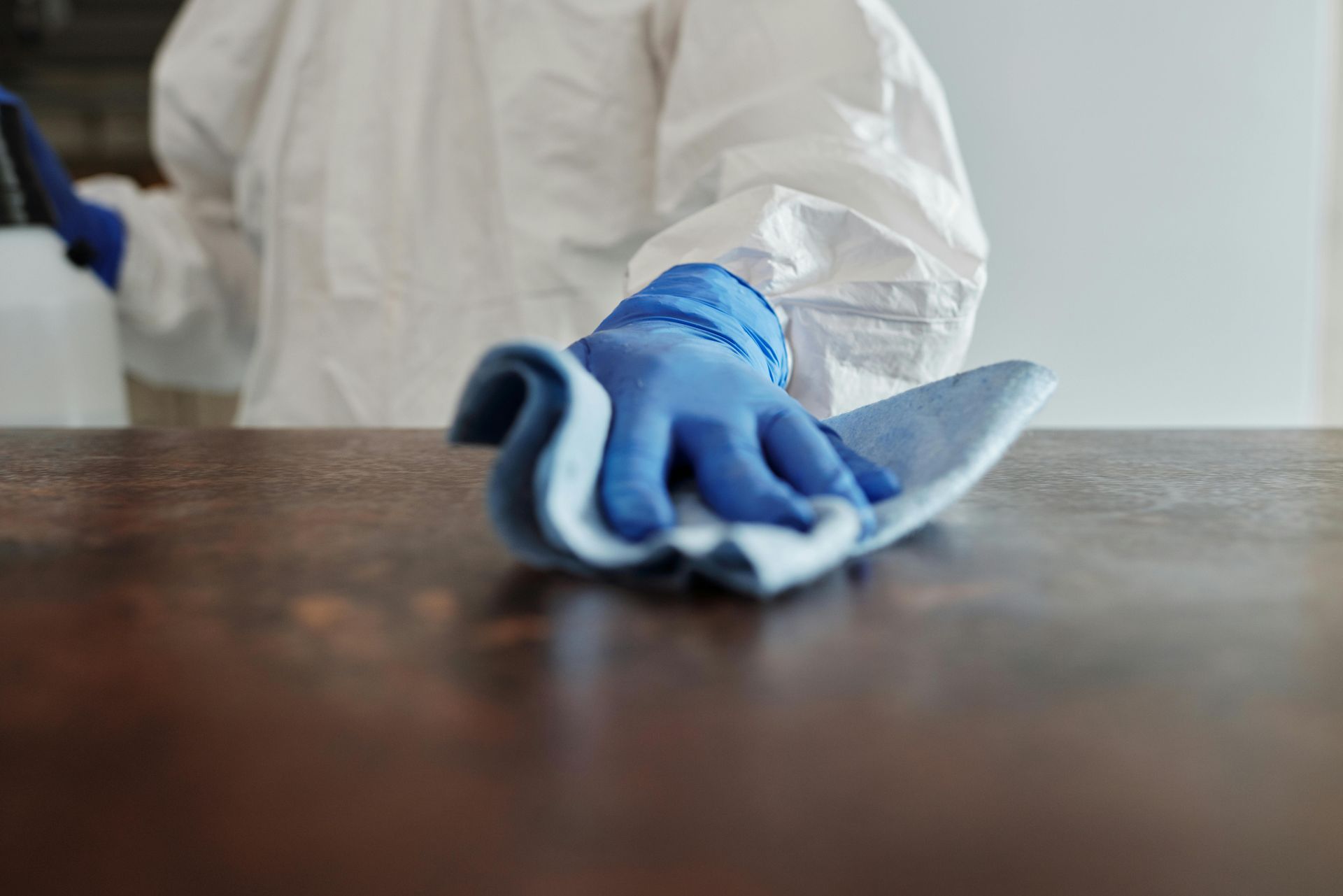 Person in a white hazmat suit and blue gloves wiping a dark wooden surface with a blue cloth.