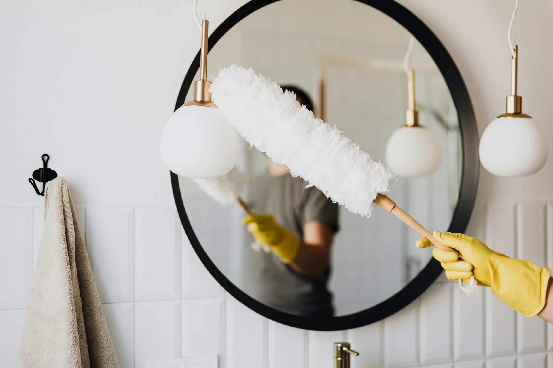 A person wearing yellow gloves dusts a round mirror in a bathroom with a fluffy duster.