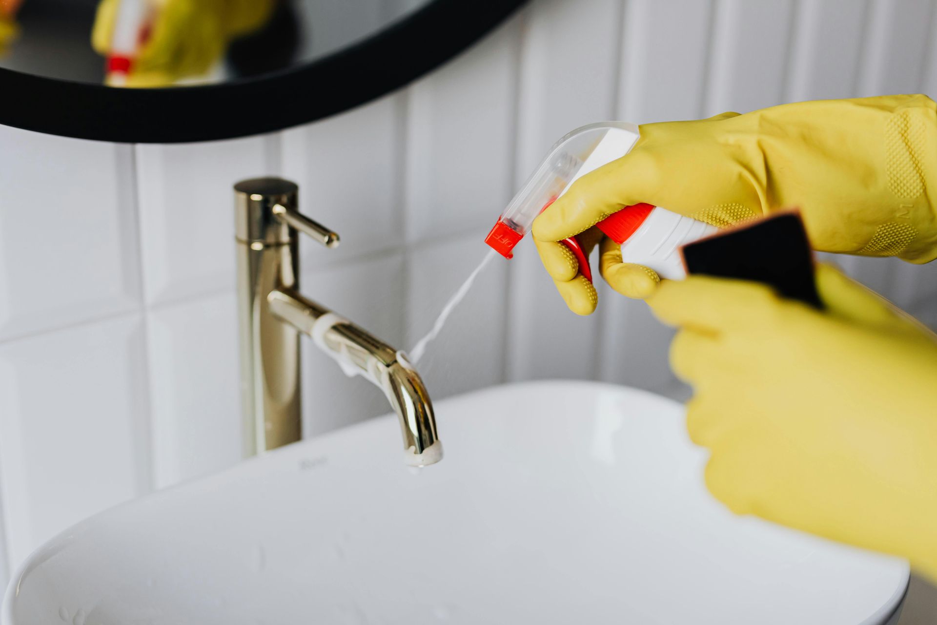 Gloved hands spray cleaner near a bathroom sink and faucet