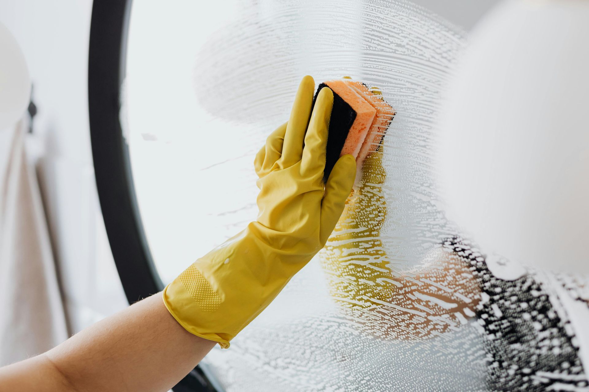 A person wearing yellow gloves dusts a round mirror in a bathroom with a fluffy duster.