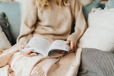 A person in a beige sweater sits on a couch under a blanket, reading an open book.