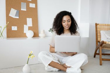 A person with dark curly hair sits cross-legged on the floor, using a laptop in a bright, minimalist home office.