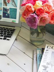 A laptop, a vase of colorful roses, and a stack of magazines on a white wooden table.