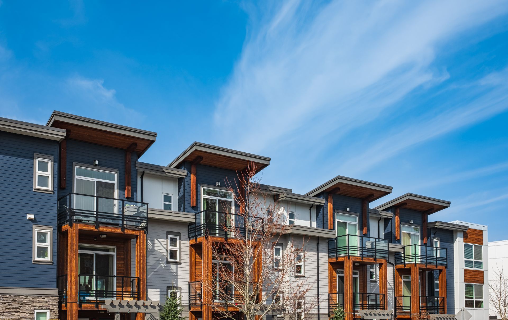 A modern multi-story apartment complex with wooden accents, dark grey siding, and balconies against a bright blue sky.