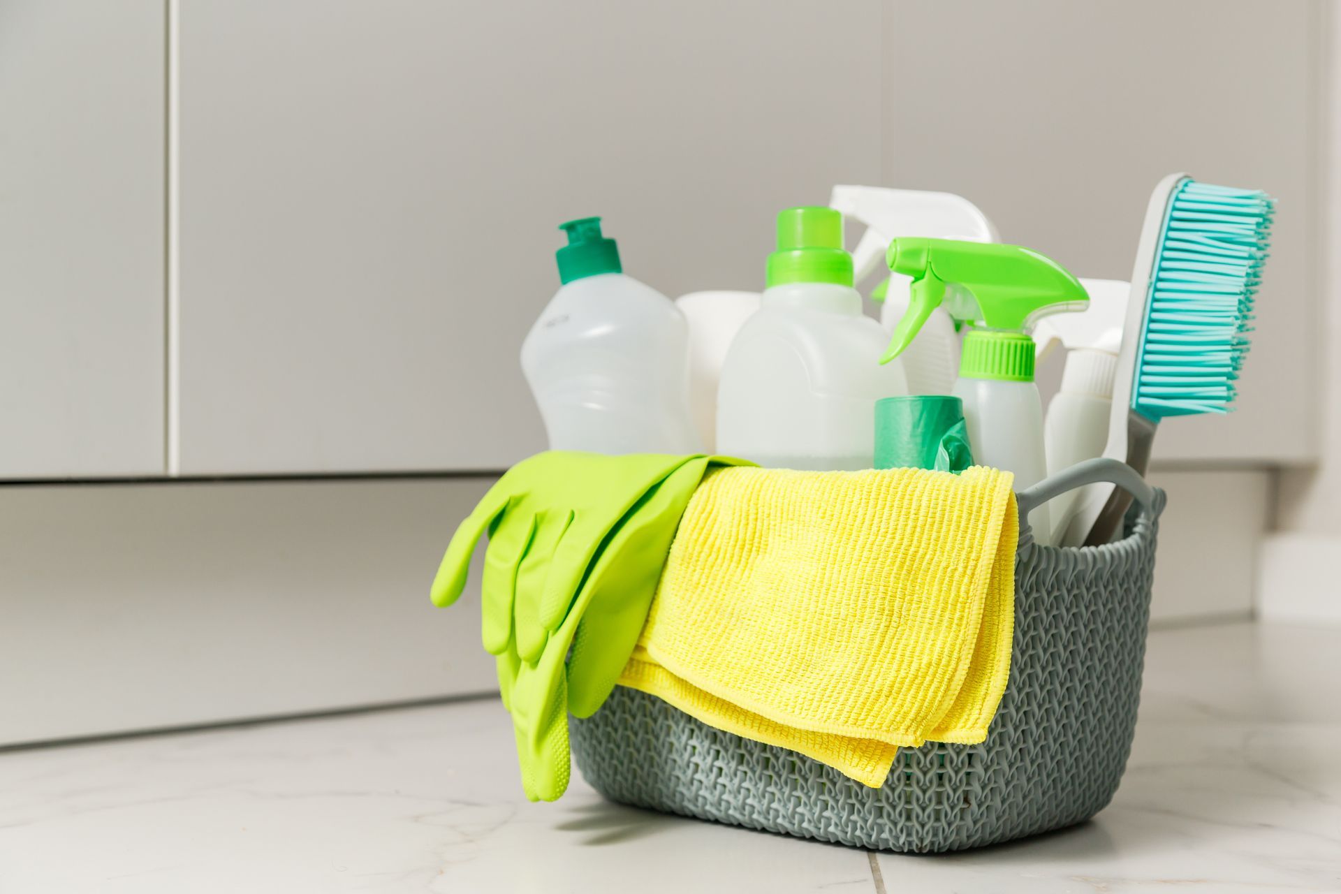 A grey woven basket filled with green cleaning supplies, including gloves, bottles, a yellow cloth, and a scrubbing brush.